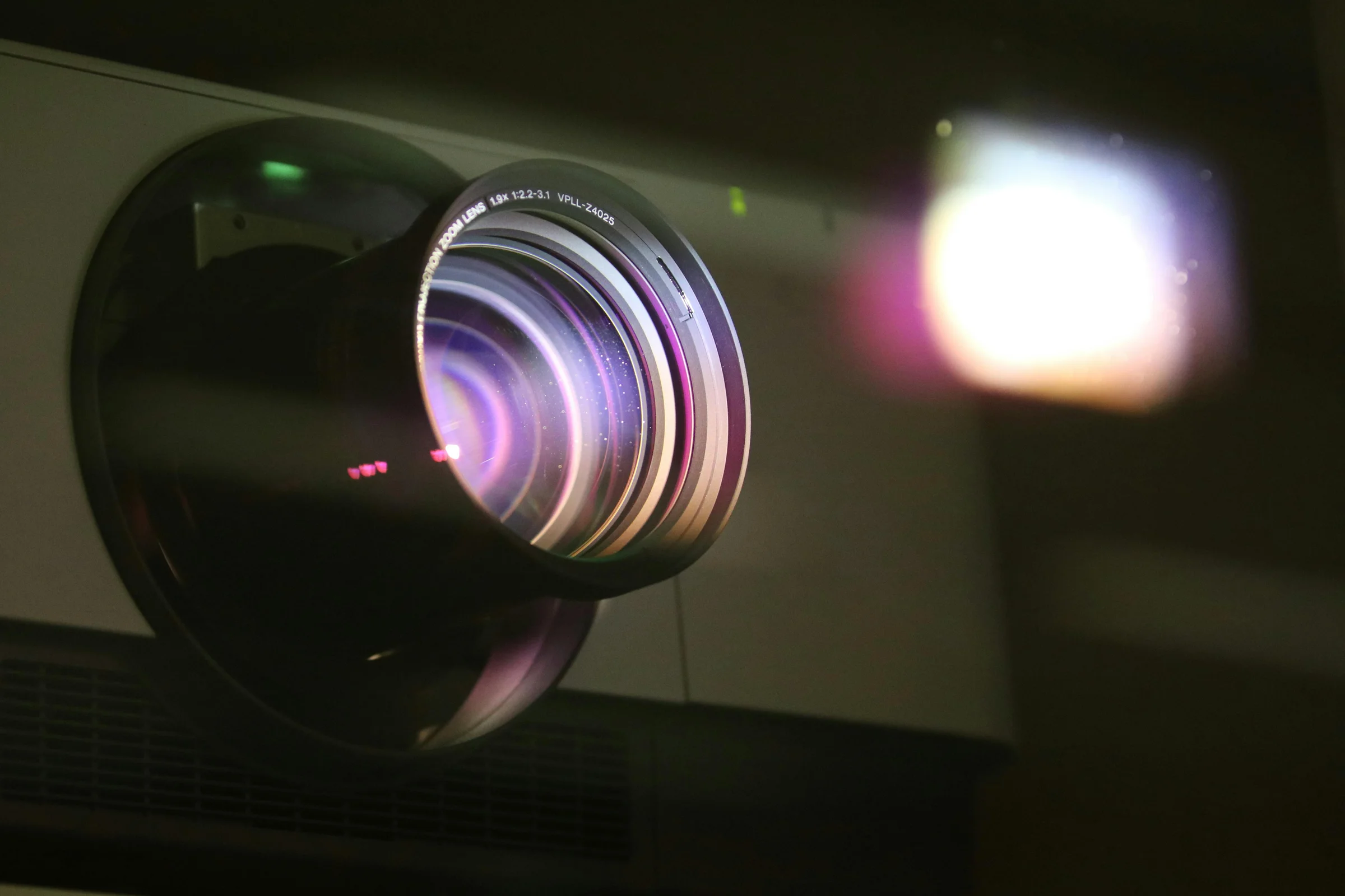Close-up of a projector lens with colorful reflections, projecting a bright image onto a dark background.